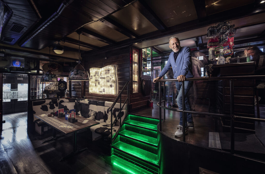 Henk de Vries standing proudly inside The Bulldog coffeeshop, surrounded by vintage décor, neon green lighting, and cannabis memorabilia.
