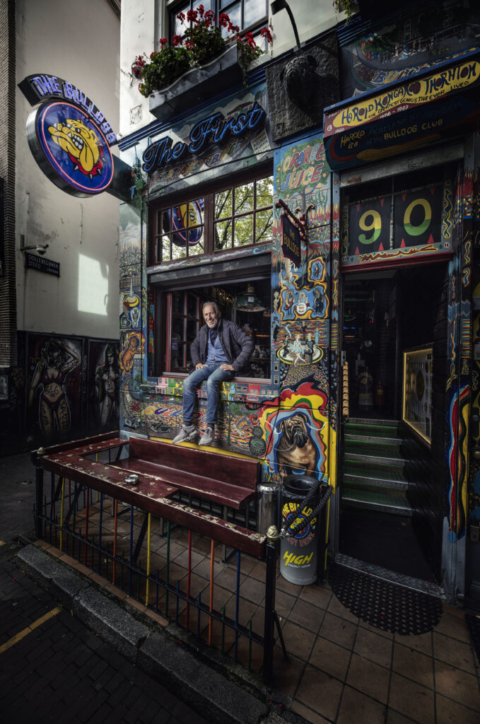 Henk de Vries seated on the windowsill outside The Bulldog’s original location, surrounded by Harold Thornton’s psychedelic street art.