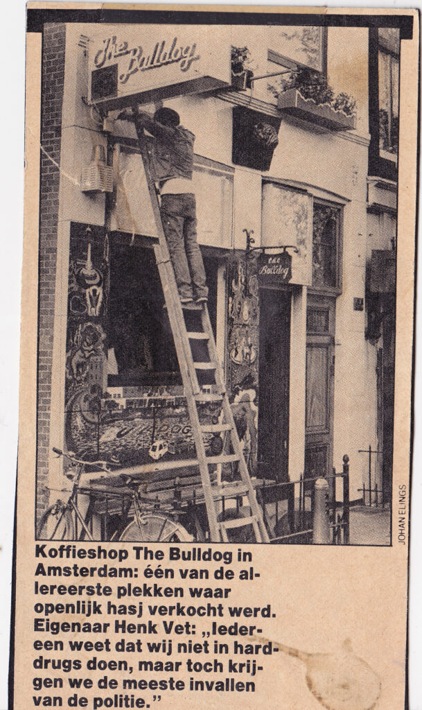 The Bulldog changed amsterdam: Black-and-white newspaper photo of someone installing The Bulldog sign above the coffeeshop entrance, with mural and bicycle visible.