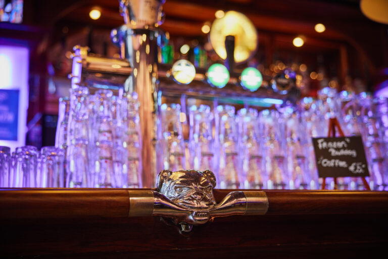 Close-up of The Bulldog bar counter handle with glassware and softly lit bottles in the background. Image used for why does cannabis sometimes smell like beer?