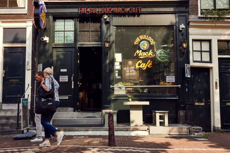Exterior of The Bulldog Mack Café in Amsterdam with black frontage, window signs and people walking past. Image used for why does cannabis sometimes smell like beer?